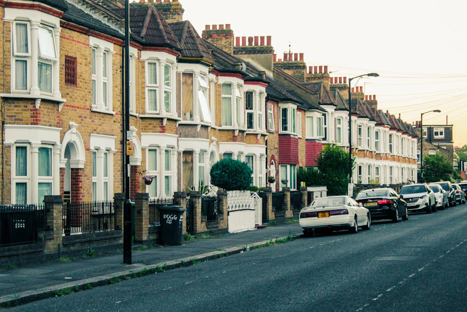 A residential street in Barking and Dagenham showing a row of terraced houses with brick facades, some with white bay windows and decorative cornices. The pavement in front of the homes features black metal fences, small garden areas with shrubs, and a black wheelie bin. Parked along the street are several cars, including a white sedan closest to the camera and darker vehicles further back. The scene is lit by soft natural light, indicating early evening or late afternoon. This setting reflects typical house exterior details relevant to home relocation and moving logistics, as part of the process managed by Man with Van Creekmouth, which offers professional removals services in the area.