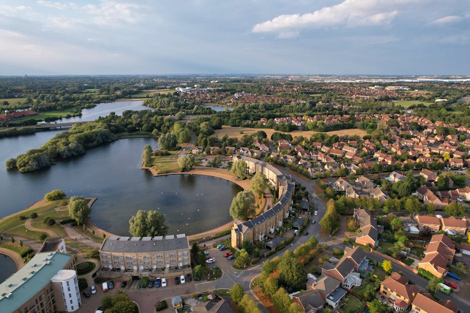 An aerial view of a residential area in Barking and Dagenham, showing a large man-made circular lake with surrounding green spaces and small trees. The lake is partially enclosed by a curved row of terraced houses with grey roofs, situated along a street where vehicles are parked. Some boats are visible on the water, and the area includes pathways with trees and benches. Adjacent to the residential houses are additional streets with more houses, gardens, and trees, extending into a suburban landscape with other water bodies and green spaces in the background under partly cloudy skies. This image exemplifies typical urban home relocation surroundings, highlighting the organized layout of houses, roads, and natural features, relevant to moving and transportation services provided by Man with Van Creekmouth for house removals in the area.