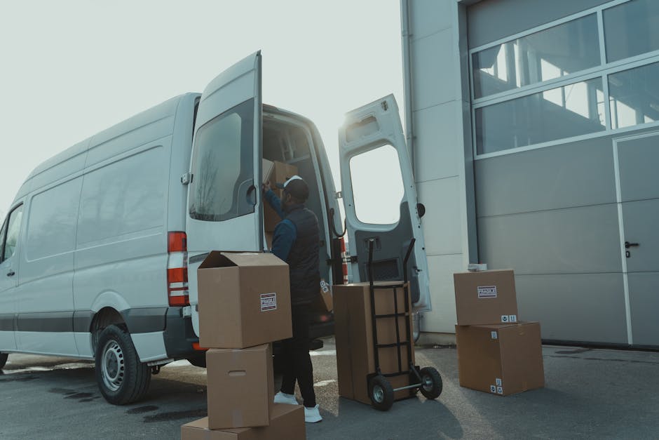 A man wearing casual clothing, including a white t-shirt, jeans, white sneakers, and a black cap, is seen loading three medium-sized cardboard boxes onto a white panel van parked on a residential stre