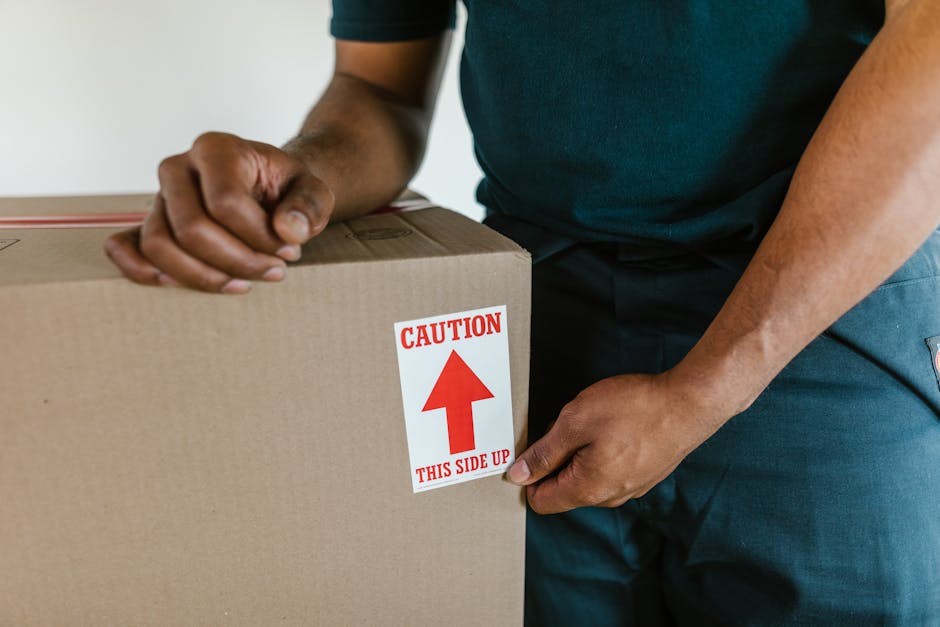 A person wearing a purple top and orange trousers is holding an open cardboard box labeled 'Fragile' with red and white handling instructions, inside a storage or packing area surrounded by several ot