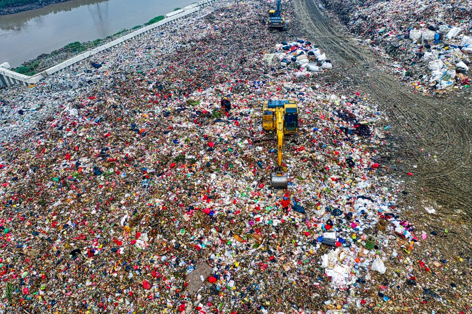 An aerial view of a large landfill site filled with a vast expanse of mixed waste, including plastic bottles, bags, cardboard, and other household debris, scattered across the uneven ground. A yellow excavator with a long arm and bucket attachment is visible in the center, actively moving or sorting the refuse amidst the mounds of rubbish. To the left, a section of a concrete boundary wall separates the waste area from the surrounding environment. The scene is outdoors under natural daylight, highlighting the extensive accumulation of waste typical of landfill or large-scale waste disposal sites, related to waste management and disposal services. Occasionally, the image reflects the scale and logistics involved in household waste removal, which connects to the broader context of removals and recycling rules, as seen on the Creekmouth movers' page. Man with Van Creekmouth specializes in responsible waste disposal and house clearance, ensuring proper handling of household items during relocation.