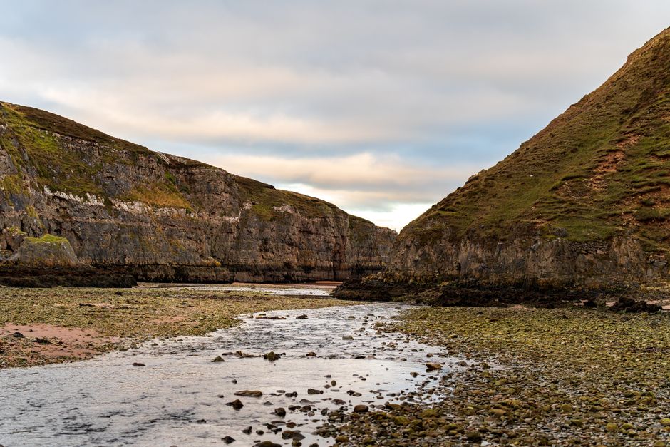 A scenic view of a narrow river or stream flowing between two steep, grassy hills with rocky cliffs. The water appears shallow with visible rocks and pebbles along the riverbed. The area surrounding the water features patches of moss and algae on the rocks, indicating a natural landscape. The sky above is partly cloudy, providing soft, diffused lighting that highlights the textures of the cliffs and vegetation. This outdoor environment is away from urban settings and not related to house removals or moving services, but it exemplifies natural terrain that could be encountered during a rural or logistical movement route.