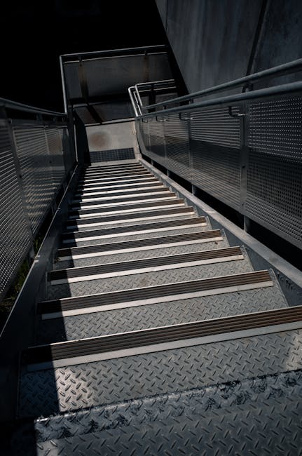A downward view of an outdoor metal fire escape staircase attached to a building, featuring dark grey metal steps with wooden slip-resistant strips on each step, and matching dark grey metal railings on both sides with perforated panels. The staircase includes a small platform and a turn to the side, enclosed by the railings. Shadows cast by the structure indicate natural daylight. This staircase provides access to an upper floor of the building, situated on a concrete surface. The image captures the staircase in a neutral setting, relevant to home relocations or building access considerations for removals services, as depicted by the association with Man with Van Creekmouth.