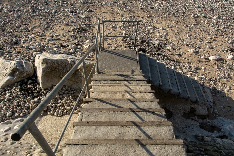 A downward view of an outdoor metal fire escape staircase attached to a building, featuring dark grey metal steps with wooden slip-resistant strips on each step, and matching dark grey metal railings on both sides with perforated panels. The staircase includes a small platform and a turn to the side, enclosed by the railings. Shadows cast by the structure indicate natural daylight. This staircase provides access to an upper floor of the building, situated on a concrete surface. The image captures the staircase in a neutral setting, relevant to home relocations or building access considerations for removals services, as depicted by the association with Man with Van Creekmouth.