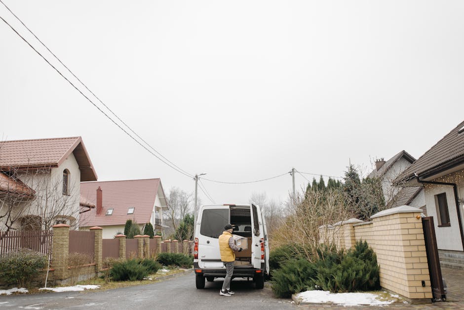 A person dressed in a yellow jacket and grey trousers is loading a cardboard box into the rear of a white van parked on a residential street, surrounded by suburban houses with pitched roofs and brick