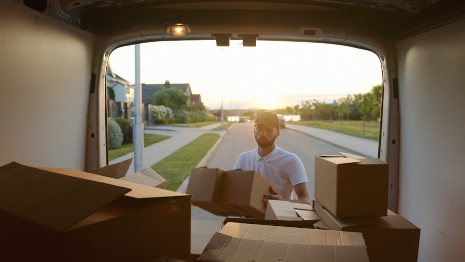A person dressed in a yellow jacket and grey trousers is loading a cardboard box into the rear of a white van parked on a residential street, surrounded by suburban houses with pitched roofs and brick