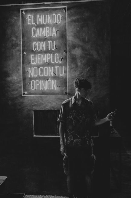 A person standing indoors near a wall and a small window, holding a smartphone in one hand. The person appears to be wearing a patterned shirt and has short hair. Behind them is a lit neon sign mounte