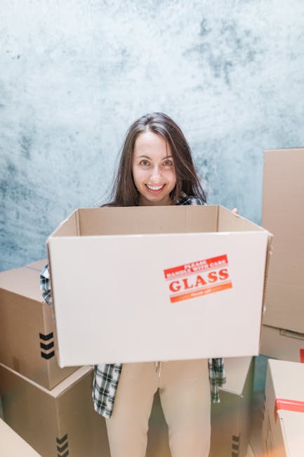 A woman with long brown hair and a checkered shirt is standing indoors against a light blue textured wall, holding a large, open cardboard box with a red 'GLASS' warning label on the side. She is smiling at the camera, and around her are several other packed cardboard boxes of various sizes, some stacked and some placed on the floor, ready for transportation. The scene depicts the process of packing and preparing household items for home relocation, as part of a furniture transport or moving service. The setting suggests an organized packing environment within a property, with a focus on fragile and non-fragile items being packed for a move, supported by the context of complete packing during a house move, which aligns with the services offered by Man with Van Creekmouth for urgent same-day removals in Creekmouth.